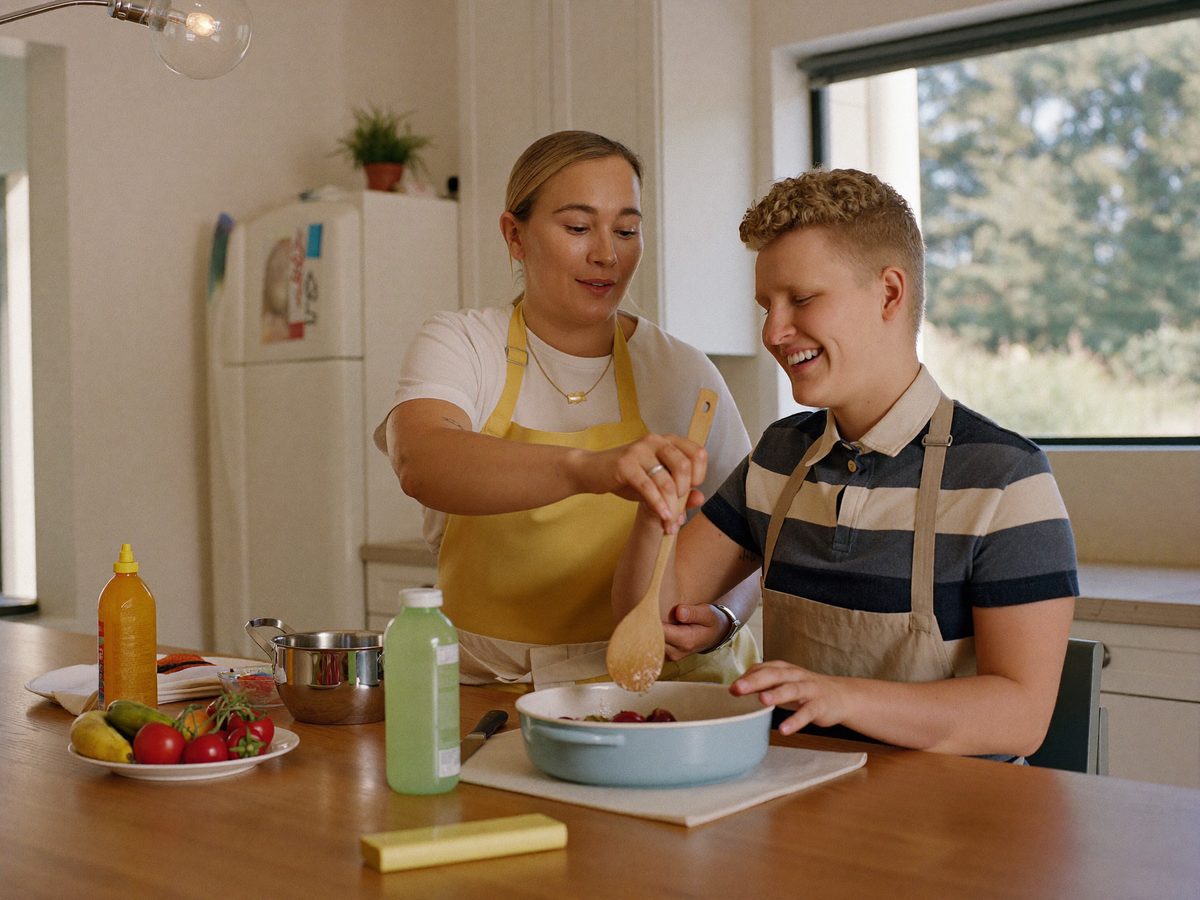 A caregiver helping a smiling young adult learn to cook in a bright kitchen.